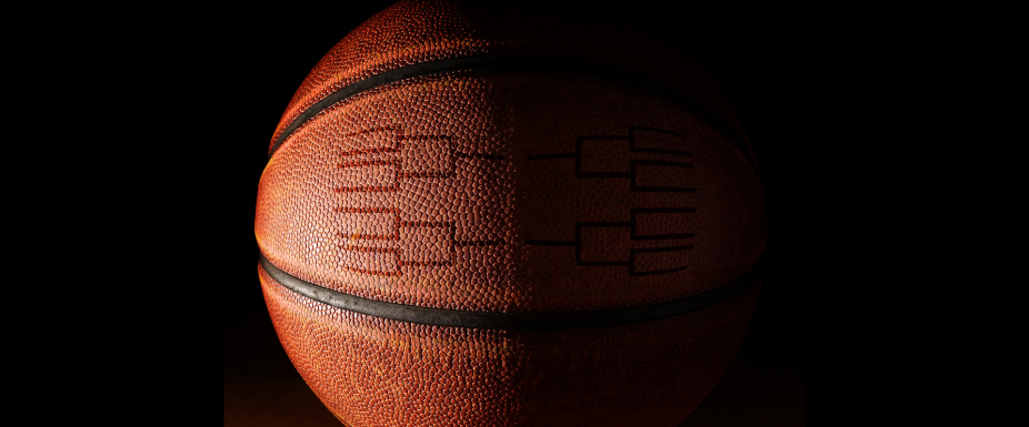 Close-up of a basketball against a dark black background, lit dramatically to highlight its textured surface and black tournament bracket graphic printed on the ball. 