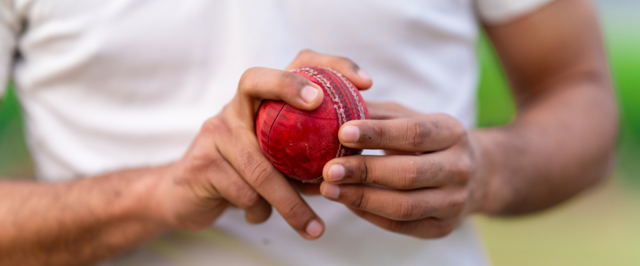 Close-up of a person’s hands gripping a worn red cricket ball, with visible stitching, against a blurred outdoor background.
