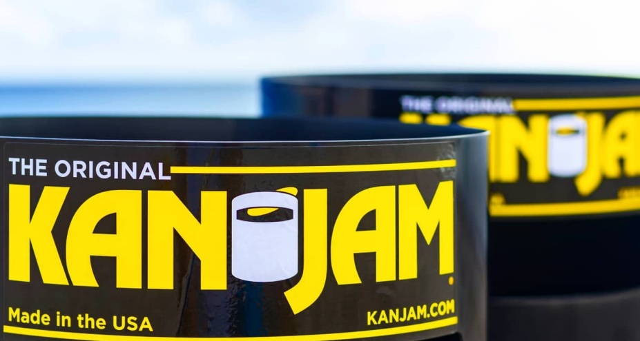 Close-up of Kan Jam game cans with bold yellow and white branding on a black background, one in focus and another blurred behind it. 