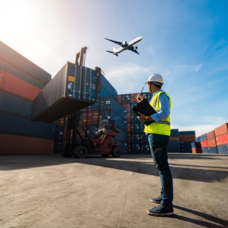 Foreman montioring shipping containers that are getting loaded to trucks while an airplane flies overhead. 