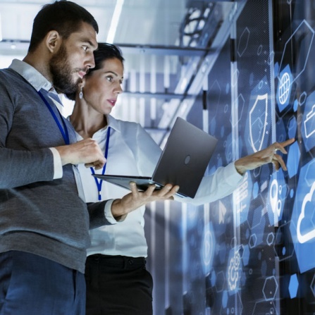 Male and female IT support associates monitoring computer systems on a laptop while reviewing a virtual display monitor on the wall. 