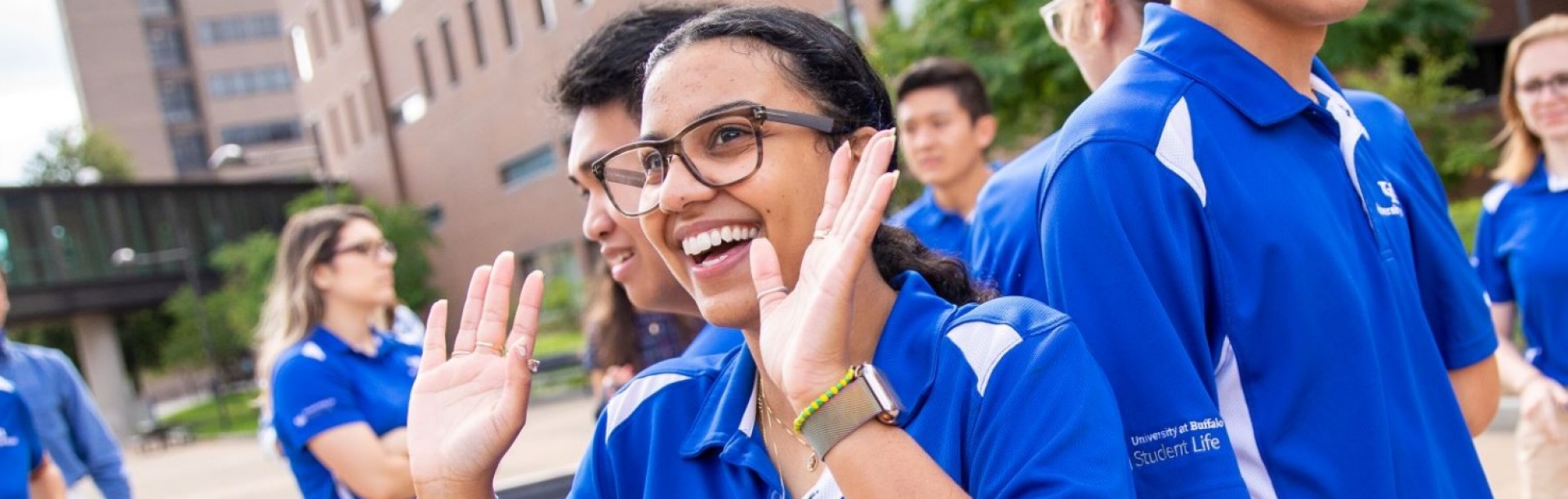 Four orientation leaders stand in the middle of a group circle during a game at a summer orientation session. All orientation leaders are wearing a version of khaki pants or shorts and their signature UB blue and white polos.
