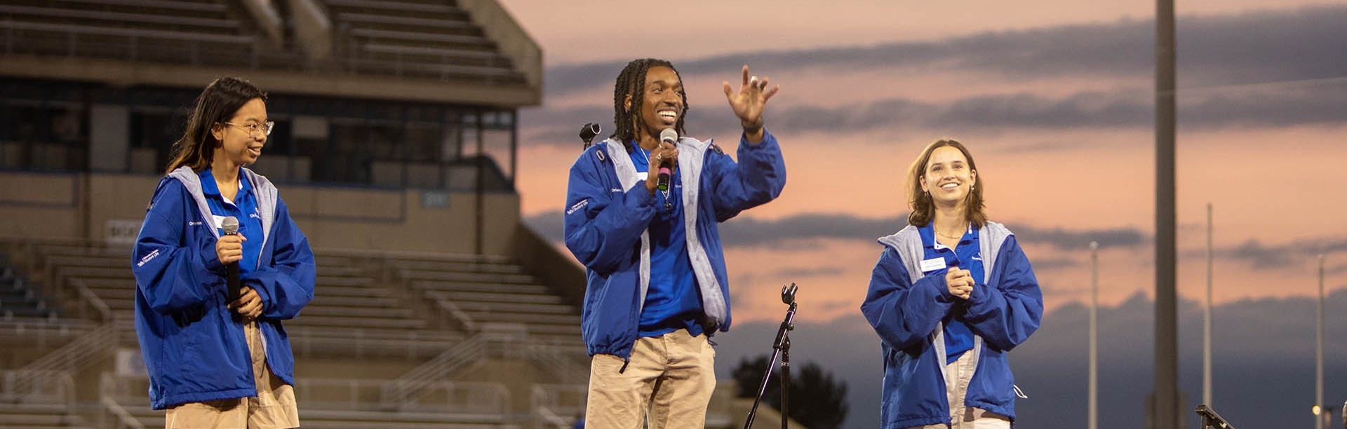 Four orientation leaders stand in the middle of a group circle during a game at a summer orientation session. All orientation leaders are wearing a version of khaki pants or shorts and their signature UB blue and white polos.