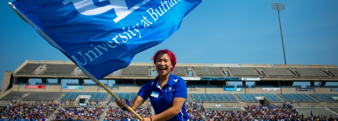 A student waves a large, blue, UB flag in front of a stadium of students at New Student Welcome. 