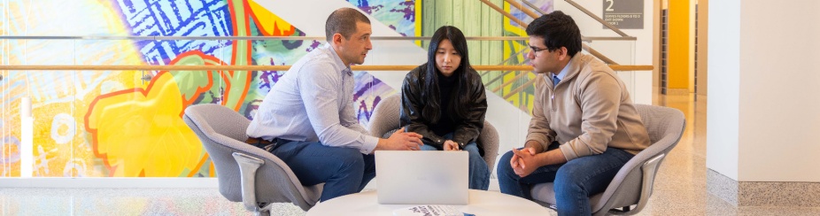 A professor sits by a round table with two students looking at a laptop. 