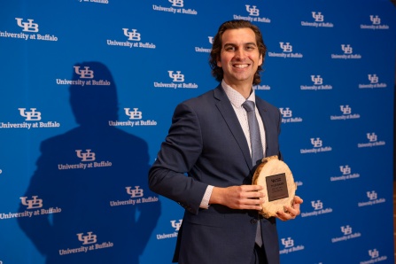 Tyler Bauer holding the 2026 SLICE award in front of a UB logo background. 