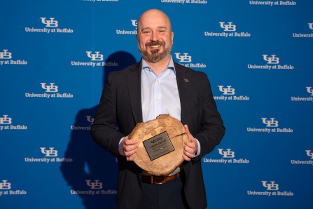 Jason Denue holding the 2026 SLICE award in front of a UB logo background. 