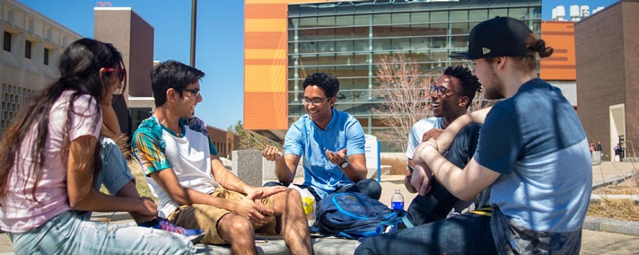 Five students sitting outside on a sunny day talking and laughing. 