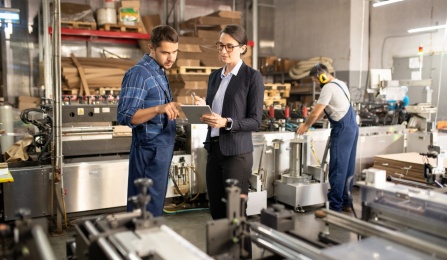 Two young workers discussing data while looking at a tablet and standing by an industrial machine. 