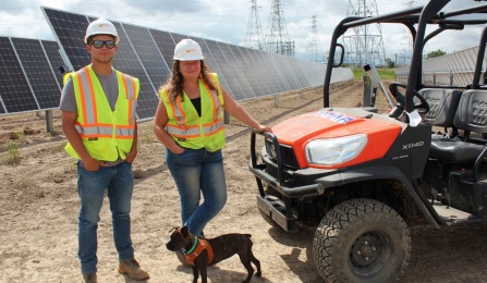 Two people wearing vests and hard hats pose at a solar array site. 