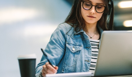 Student writing in a book while sitting at desk with laptop and coffee cup. 