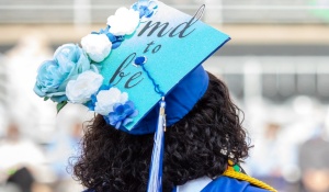 A student poses with her custom graduation cap. Coupled with light blue, royal blue, and white flowers, the cap reads "MD to be" in black letters on a teal background. 