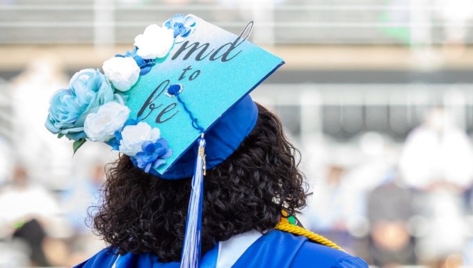 A student poses with her custom graduation cap. Coupled with light blue, royal blue, and white flowers, the cap reads "MD to be" in black letters on a teal background. 