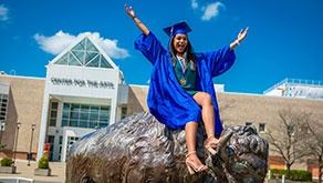 Woman in blue cap and gown smiles for a photo on top of a bronze buffalo statue. 