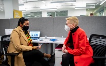 Two professionals wearing masks talking at a desk in a UB office. 