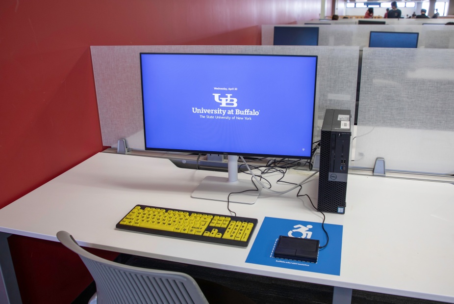 Accessible workstation at the University at Buffalo, featuring a large monitor, high-contrast yellow-on-black keyboard, assistive trackpad on an accessibility symbol mousepad, and desktop computer. 