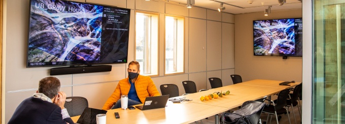 Two staff sit in a GRoW conference room with display panels on the wall and laptops open before them. 