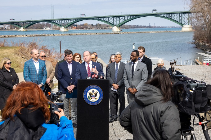 Rep. Tim Kennedy at a podium surrounded by several people, the Peace Bridge in the background. 