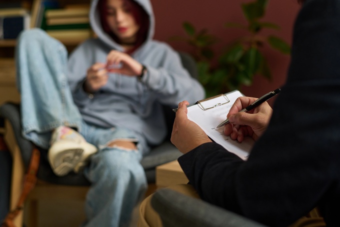 Disinterested teen lounges in a chair being interviewed by a person with a notepad. 