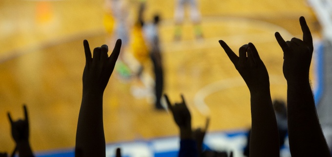 Silhouette of fans hands making a sign of the horns with a basketball court is in the background. 