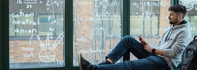 A student listening to his phone near a window covered in equations. 