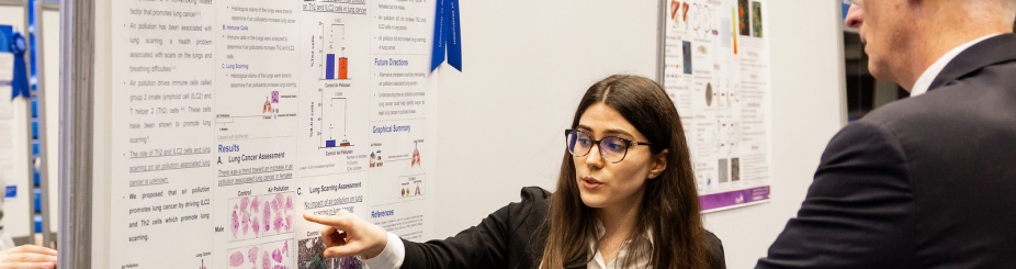 Student standing in front of her research poster points to information while Vice Provost Graham Hammill looks on. 