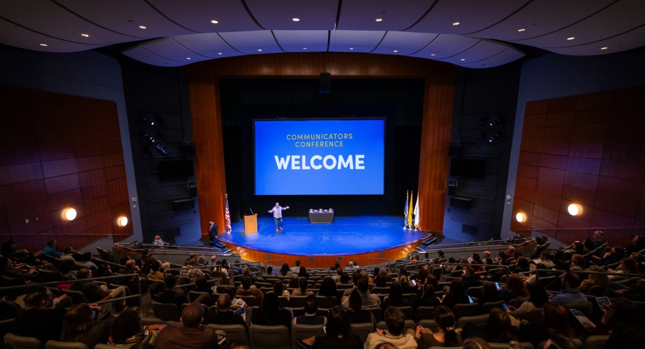 Wide auditorium filled with attendees watching a keynote presenter on a stage at the Communicators Conference. 