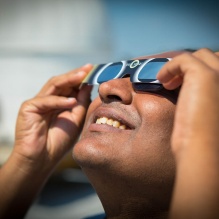 Afrincan American male wearing solar eclipse glasses while looking upwards. 