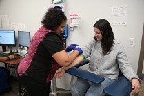 Student sitting in a chair in a medical facility, with a healthcare professional putting cloth around her arm. 